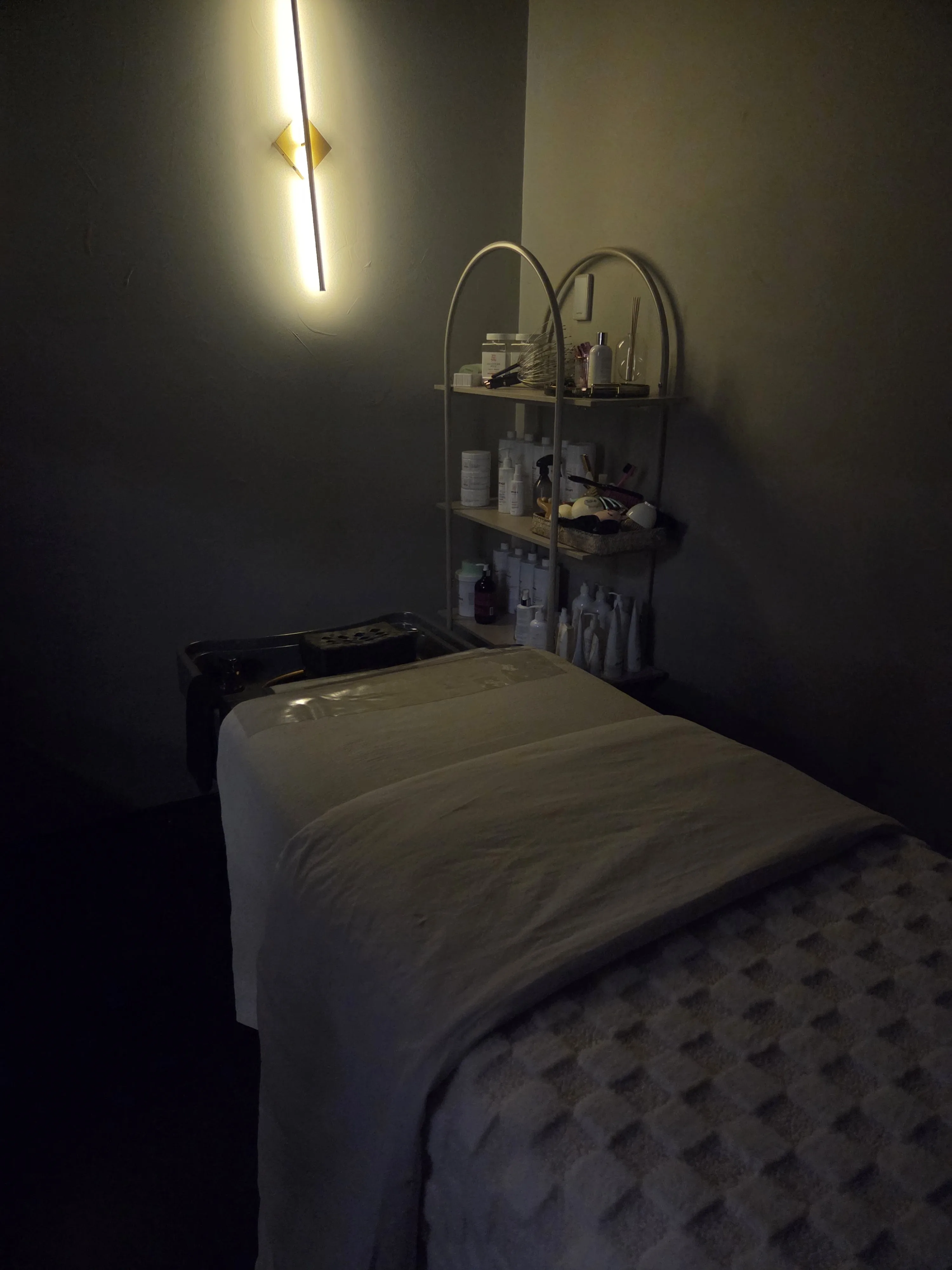 The private MIYABI Head Spa treatment room — cream-linen bed, linear brushed-gold sconce, arched product shelving, warm dark walls